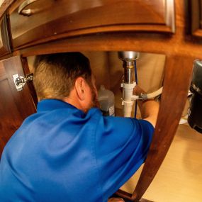 A plumber from Covenant Plumbing in the Bloomington-Normal, IL area works under a kitchen sink. The plumber, identified by their logo-emblazoned uniform, skillfully addresses plumbing issues, ensuring optimal kitchen functionality