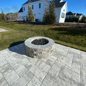 A cozy backyard in Delaware featuring a circular stone firepit surrounded by a ring of rustic wooden chairs. The firepit is lit, casting a warm glow on the faces of several people enjoying the evening. Lush greenery and trees can be seen in the softly lit background, enhancing the intimate outdoor setting.