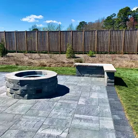 A cozy backyard in Delaware featuring a circular stone firepit surrounded by a ring of rustic wooden chairs. The firepit is lit, casting a warm glow on the faces of several people enjoying the evening. Lush greenery and trees can be seen in the softly lit background, enhancing the intimate outdoor setting.