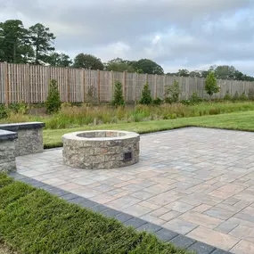 A cozy backyard in Delaware featuring a circular stone firepit surrounded by a ring of rustic wooden chairs. The firepit is lit, casting a warm glow on the faces of several people enjoying the evening. Lush greenery and trees can be seen in the softly lit background, enhancing the intimate outdoor setting.