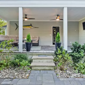 A professional hardscaping project underway in a Delaware home's backyard. The image shows landscapers laying irregular stone pavers to create a winding pathway. The scene includes a variety of landscaping tools and materials like sand and stone, with a partially completed patio area visible in the background. The house and garden display early spring foliage.