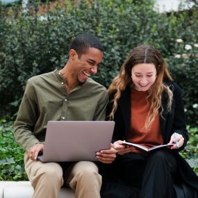 a man and woman sitting on a bench looking at a laptop computer