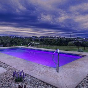 Pool with purple lights in a backyard surrounded by pavement and stone.