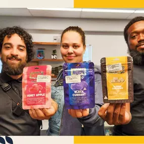 Three budtenders at The Living Room Dispensary in Maryland smiling and holding up cannabis edibles in flavors Honey Apple, Black Currant, and Blonde Chocolate.