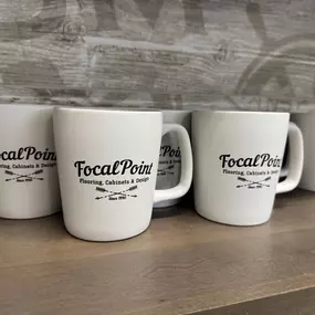 White mugs featuring the Focal Point Flooring logo and tagline. The logo includes the company name, a crossed arrow design, and the year 1990.