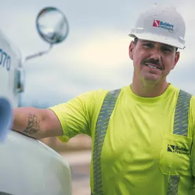 A Builders FirstSource delivery driver stands confidently next to a branded truck, wearing a white hard hat and neon yellow reflective shirt.