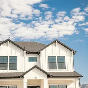 A modern two-story white house with black-framed windows stands against a bright blue sky filled with scattered white clouds. The home features clean vertical siding and sharp architectural lines, including twin gables and a centered entryway with a small overhang. The number 