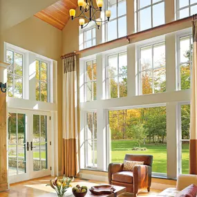 A bright and airy living room with soaring ceilings and large, floor-to-ceiling windows that let in natural light and showcase a lush green lawn and trees with autumn foliage. The space features a glass double-door entry, elegant drapes, a leather armchair, and a wooden coffee table with decorative accents.