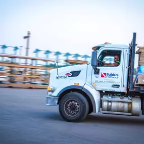 A Builders FirstSource flatbed semi-truck, stacked with lumber and construction materials, is driven by a uniformed employee through a lumberyard. This action shot emphasizes reliable material delivery, fleet operations, and construction logistics services.