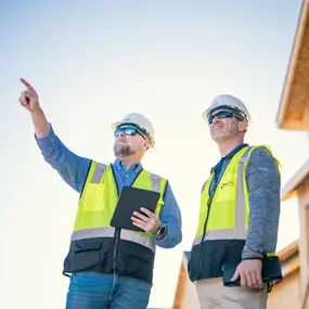 Two Builders FirstSource professionals wearing hard hats and reflective vests stand outside a framed home, reviewing plans on a tablet and pointing toward construction progress. The image highlights modern project management, jobsite planning, and teamwork in residential building.