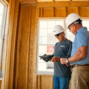 Two Builders FirstSource professionals, wearing hard hats and safety glasses, review digital construction plans on a tablet inside a framed residential home. Surrounded by exposed OSB walls and natural light, the scene highlights tech-enabled project management in new home construction.