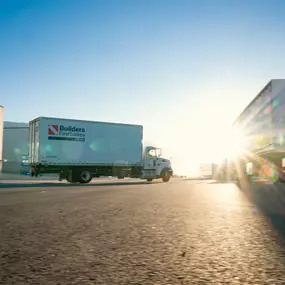 Builders FirstSource delivery trucks on the move outside a distribution facility at sunrise, symbolizing nationwide reach and early-start dedication.