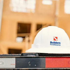 A white Builders FirstSource hard hat sits atop a flatbed truck labeled “CLARK” at a construction site. The background shows a building under construction with exposed framing and scaffolding, emphasizing jobsite safety and professionalism.