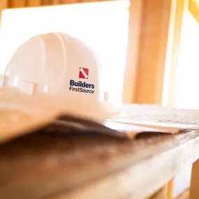 A white Builders FirstSource hard hat rests on a beam of lumber inside a partially framed home, glowing with natural sunlight through the windows. The image emphasizes construction jobsite safety, framing materials, and new residential builds