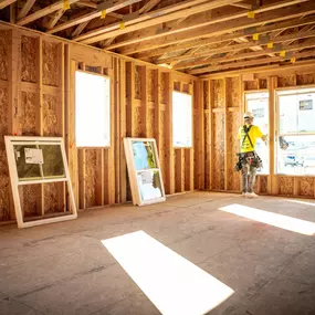 A construction worker in a bright yellow safety shirt and hard hat stands inside the wooden frame of a house under construction. The unfinished room features exposed plywood walls and ceiling beams, with large rectangular window openings. Two uninstalled windows lean against the wall, and several installed windows on the right let in natural light, casting bright rectangular patches on the floor.