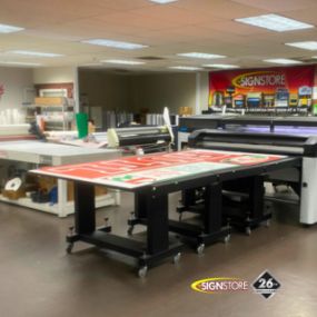 Signstore vinyl production department featuring large printing tables, printers, and a workstation with various signage materials. The area is well-organized and designed for high-volume production. The 
