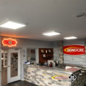 Signstore reception area with a stone counter, large Signstore logo on the wall, and wood-like flooring. An illuminated open sign is above a doorway on the left, and various certificates and awards are displayed behind the counter.