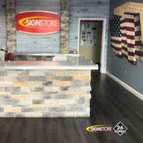 Signstore reception area featuring a stone counter and a large Signstore logo on the wall behind it. The area has a wood-like floor, and a decorative American flag is displayed on the right wall. The 