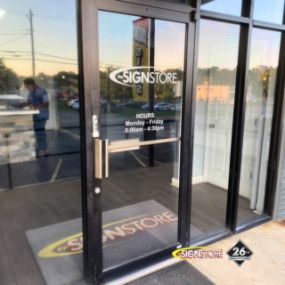 Entrance door of Signstore showing business hours: Monday - Friday, 8:00am - 4:30pm. The glass door has the Signstore logo, and the interior features a mat with the Signstore branding. The 