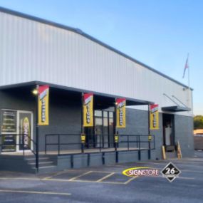 Exterior of Signstore building featuring modern white and gray design. Vertical banners read “SIGNSTORE” and “SIGN CENTER.” The entrance has a ramp and stairs. Signstore logo and 