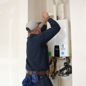 A picture of a man standing in front of a tankless water heater holding a tool repairing something