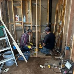 A picture of men working on a wall with exposed studs, water lines and electrical lines.