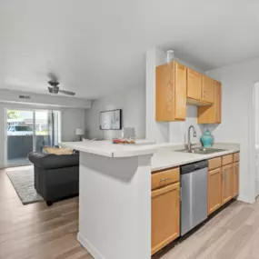 View from kitchen with plank flooring, wooden cabintery, breakfast bar, sink, and dishwasher into the living area