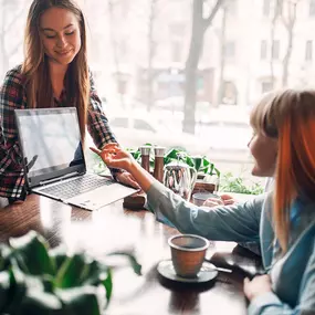 Zwei Frauen sitzen in einem Café, während eine von ihnen auf einen Laptop zeigt, der auf dem Tisch steht. Das Bild zeigt ein informelles Geschäftstreffen oder eine Beratungssituation, im Zusammenhang mit gastronovi-Dienstleistungen, bei denen digitale Lösungen wie zum Beispiel ein Software-Modul für die Warenwirtschaft, in der Gastronomie diskutiert werden.