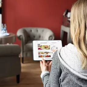 Eine Frau sitzt auf einem Sofa in einem geschmackvoll eingerichteten Raum und navigiert auf ihrem Tablet durch eine digitale Speisekarte von gastronovi. Dieses Bild zeigt den praktischen Einsatz digitaler Lösungen zur Erhöhung des Komforts für Restaurantbesucher. Die Szene vermittelt ein modernes Kundenerlebnis in der Gastronomie, bei dem Gäste bequem Bestellungen aufgeben können. Durch das Bestellsystem von gastronovi können Gäste Bestellungen mobil aufgeben und auf Wunsch direkt online bezahle