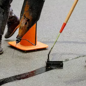 Workers are applying hot sealant to cracks in the asphalt. One worker is handling a piece of equipment that dispenses the sealant, while another uses a tool to smooth and guide the sealant into the crack.