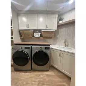 This remodeled laundry room features light-colored cabinetry, coordinated countertops and flooring, and a new washer and dryer set. A custom overhead storage area above the machines maximizes functionality, bringing clean design and smart organization to this updated utility space.