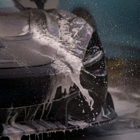 A close-up of a silver car being washed with a soapy sponge. The car is parked in a garage or bay with a concrete floor. Water is dripping down the side of the car.