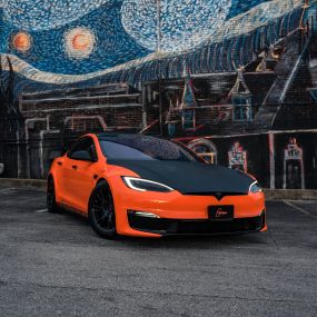 A bright orange Tesla Model S electric car is parked in front of a colorful mural. The mural depicts a variety of shapes and figures in orange, blue, and yellow. The Tesla has a sleek, futuristic design with a panoramic glass roof and chrome trim.