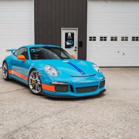 A bright blue and orange Porsche 911 GT3 RS wrapped by CWRAPS parked in front of a closed white garage door. The car has a large black rear wing and black wheels with orange rims. The text 