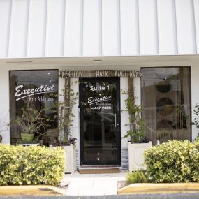 The front entrance of Executive Hair Additions in Boca Raton, FL, features a white awning and glass door with the business name and contact information. The entrance is adorned with green shrubbery, creating an inviting atmosphere for clients.