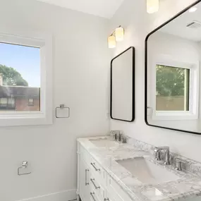 Bright and inviting bathroom design with a double-sink vanity, dual mirrors, and chic modern lighting. The large window floods the space with natural daylight, highlighting the fresh white walls and shower. A perfect blend of style and functionality!