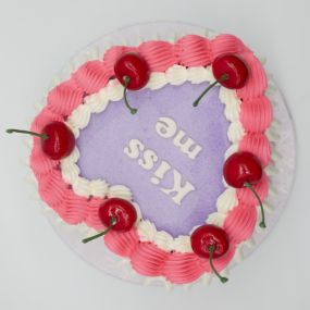 Heart cake with purple frosting, pink and white frosting border, and topped with cherries. The cake features the message 