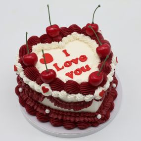 A tall heart cake with white frosting, decorated with red and white frosting borders, and topped with cherries. The cake features the message 
