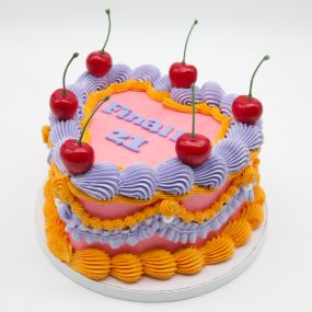 A tall heart cake with layers of bright pink frosting, accented with intricate swirls of purple and yellow frosting. The cake is topped with red cherries and features the message 