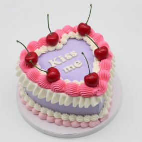 A tall heart cake with purple frosting, decorated with pink and white frosting borders and topped with cherries. The cake features the message 