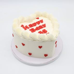 A tall heart cake with smooth white frosting, decorated with small red hearts along the sides. The top features the message 