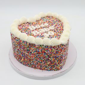 A tall, heart-shaped cake adorned with colorful sprinkles on the sides and top. The top of the cake features the message 