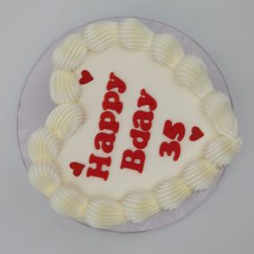 A heart-shaped cake with smooth white frosting, decorated with swirls of white frosting around the edge. The cake features the message 