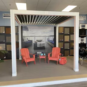 A showroom display featuring a stylish outdoor living setup with composite decking, a modern pergola, and vibrant chairs. The backdrop showcases a scenic waterfront view, while wall-mounted decking samples highlight available colors and materials. Ideal for customers exploring premium decking and millwork solutions.