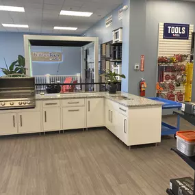 A modern outdoor kitchen display at Builders FirstSource featuring a stainless steel grill, white cabinetry with granite countertops, and a TimberTech pergola. The showroom highlights decking materials, millwork, and tool selection for home improvement and outdoor living projects.