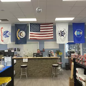 The front counter at Builders FirstSource Chesapeake location, adorned with multiple American and military service flags. The customer service desk is stocked with essential building materials, fasteners, and hardware supplies, reflecting a commitment to quality construction and support for service members.