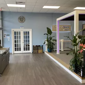 A showroom display featuring TimberTech composite decking with an outdoor pergola setup. The space is decorated with lush green plants, vibrant chairs, and a modern grilling station, showcasing premium decking and outdoor living solutions.
