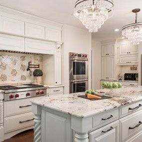 Elegant kitchen remodel featuring white kitchen cabinets, a central island with a light blue base, and crystal chandeliers. Stainless steel appliances and a decorative backsplash complete the luxurious look. Photo by Deanna Bert.