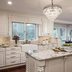 This elegant kitchen remodel showcases classic white kitchen cabinets paired with luxurious marble countertops and ornate chandeliers. The natural stone backsplash adds a rustic charm to the sophisticated design. Photo by Deanna Bert