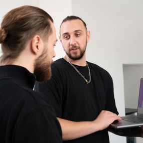 Zwei bärtige Männer in schwarzen Shirts diskutieren vor einem Laptop. Einer trägt einen Dutt und ist im Profil zu sehen, der andere mit Kette blickt in die Kamera. Sie wirken konzentriert und engagiert in ihrer Zusammenarbeit.